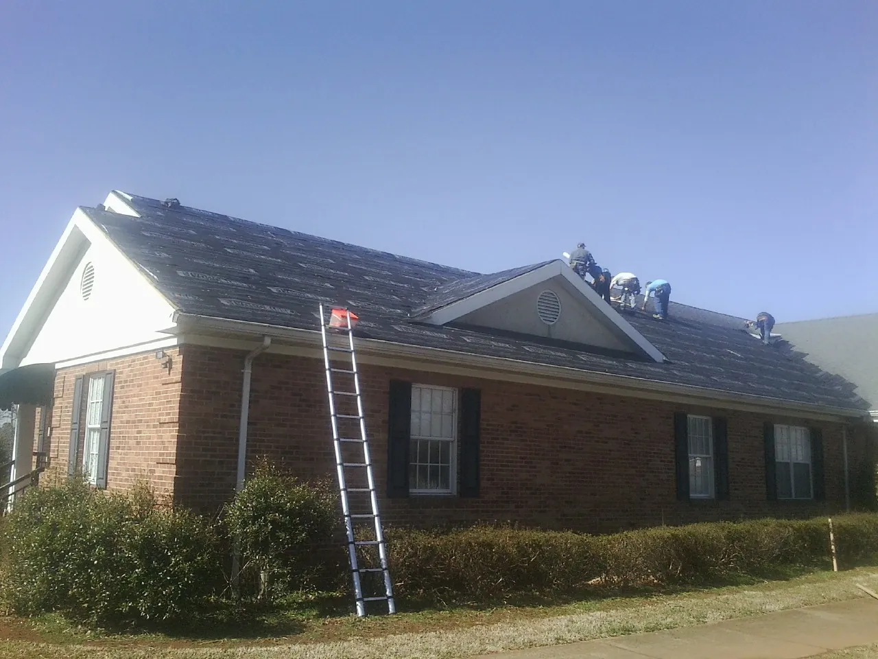 Crew tearing shingles off a brick ranch roof with a ladder set — Emergency Roof Repair work by our crew in Wanamingo