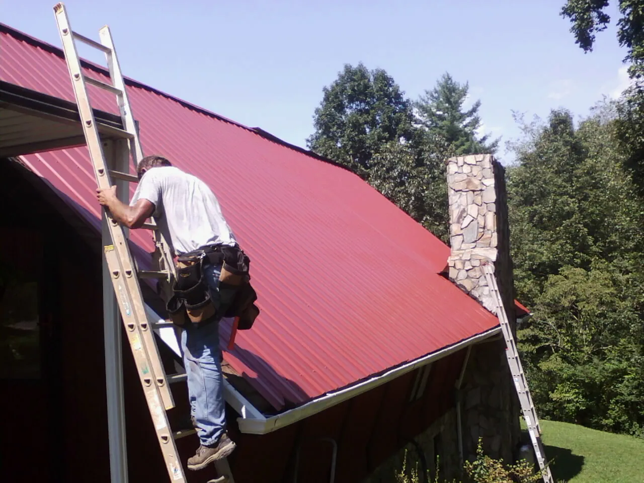 Roofer on a ladder beside a red metal roof and stone chimney — Soffit & Fascia Repair by Roofing Brothers in Wanamingo